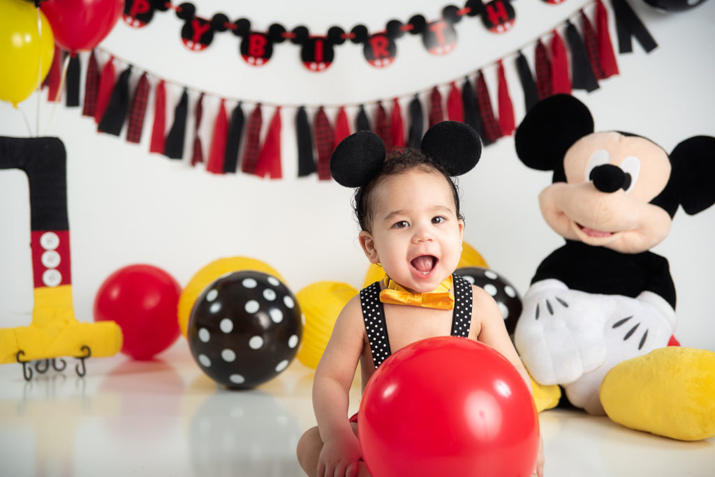 Mickey Mouse-inspired first birthday photoshoot with baby girl, balloons, and large stuffed Mickey prop
