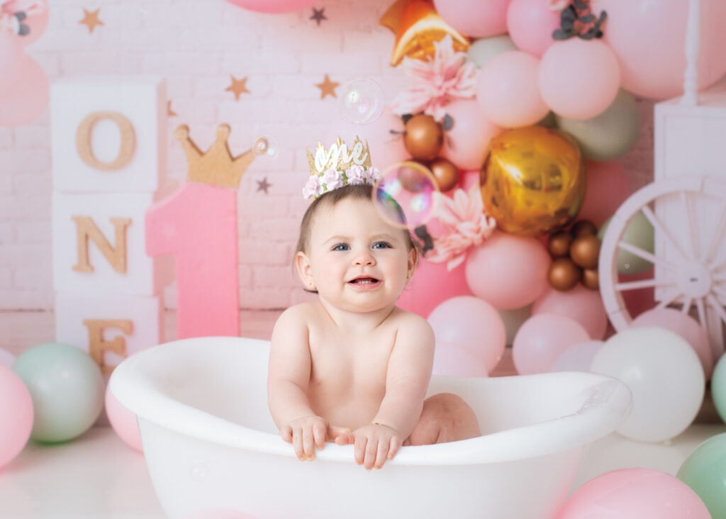 Baby girl smiling at bubble in white clawfoot tub after her princess cake smash