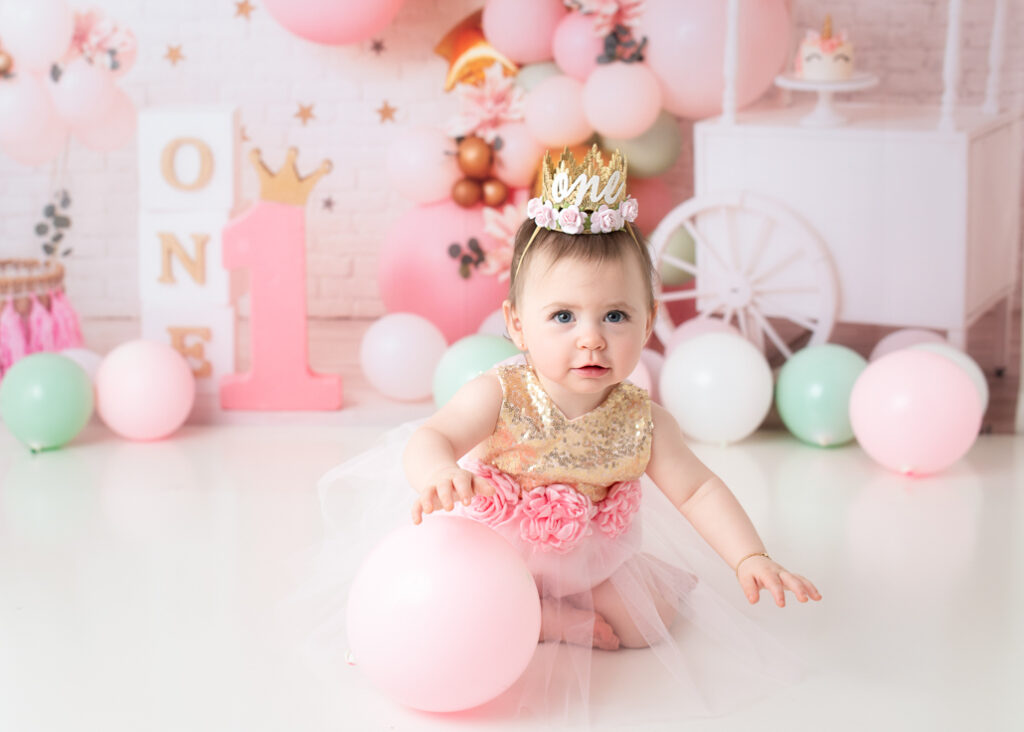 Birthday girl looking up and crawling while holding a pink balloon, wearing a crown and tulle skirt