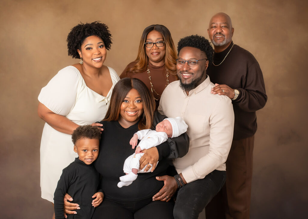 Family with one toddler and newborn and an Aunt, Grandma, and Grandpa posed in front of a tan textured backdrop.