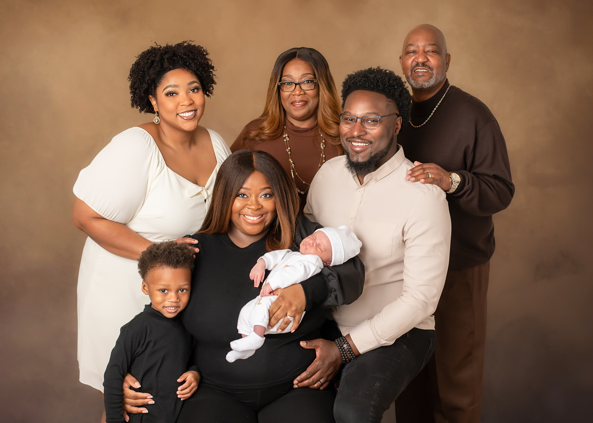 Family with one toddler and newborn and an Aunt, Grandma, and Grandpa posed in front of a tan textured backdrop.
