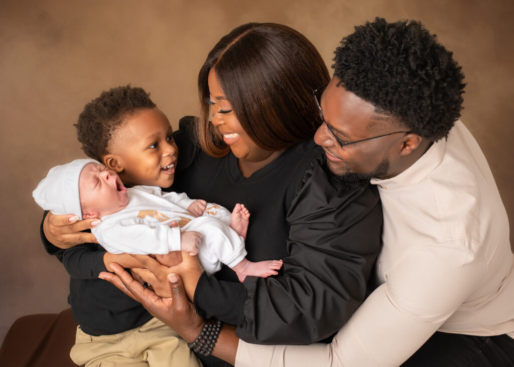 Mother and father smiling as their toddler lovingly gazes at his newborn baby sibling, all sharing a joyful family moment in a studio portrait.