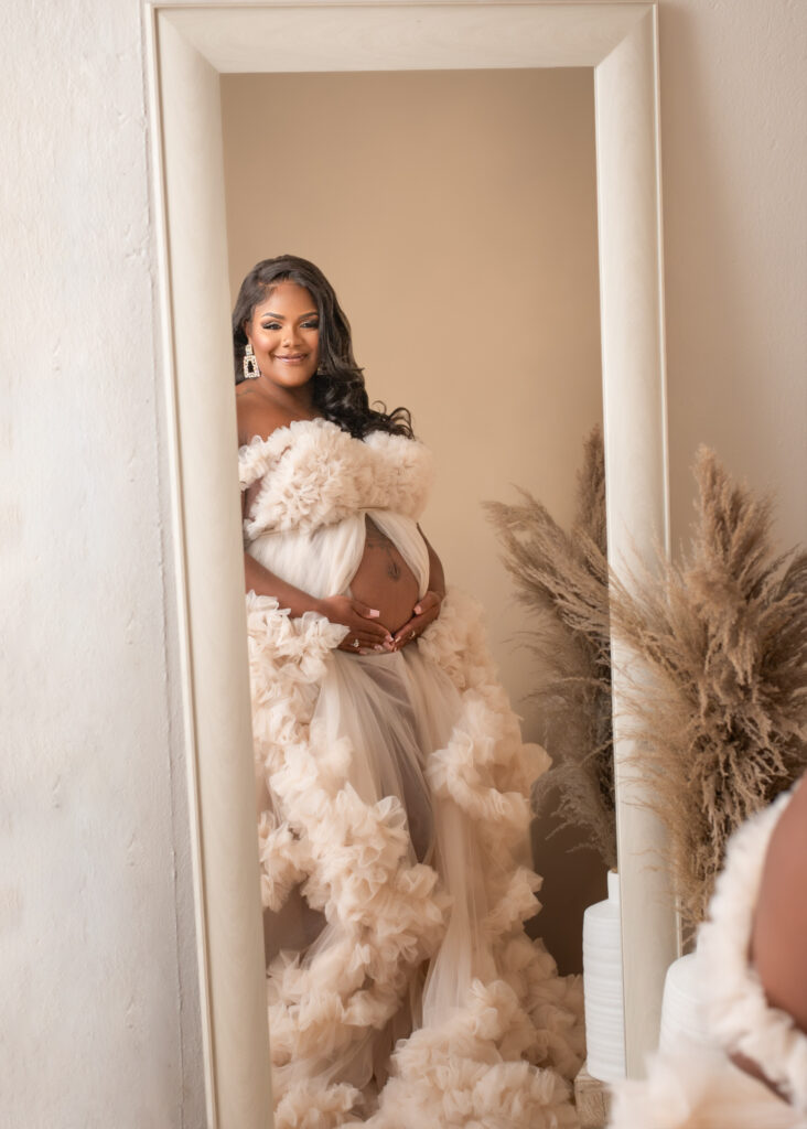 Pregnant woman in a dramatic tulle gown poses in front of a mirror, gently holding her belly and smiling, surrounded by soft neutral tones and pampas grass.
