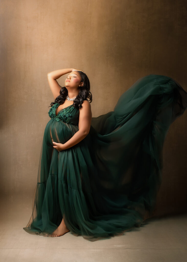 Fine art maternity portrait of a pregnant woman standing barefoot against a neutral studio backdrop, wearing a dark green flowing gown with delicate lace bodice, one hand cradling her belly and the other resting on her head as the fabric billows gracefully behind her.