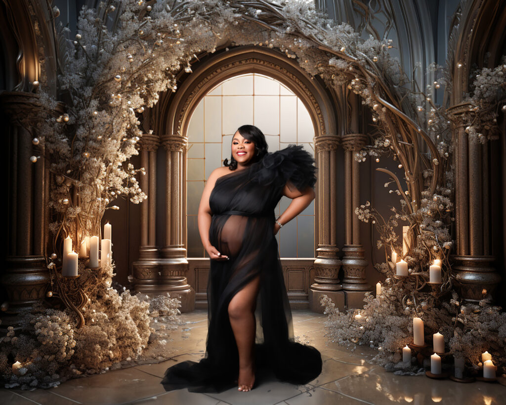 Fine art maternity portrait of a pregnant woman standing barefoot in an ornate, candlelit archway. She wears a sheer black one‑shoulder gown with a dramatic tulle sleeve, one hand cradling her belly and the other resting on her hip, surrounded by pillar candles and delicate floral arrangements against a richly textured backdrop.