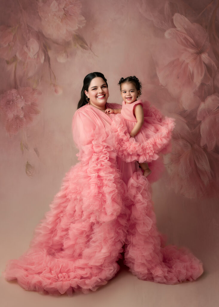 Studio portrait of a smiling mother holding her young daughter; both dressed in matching voluminous pink tulle gowns against a soft, painterly floral backdrop.