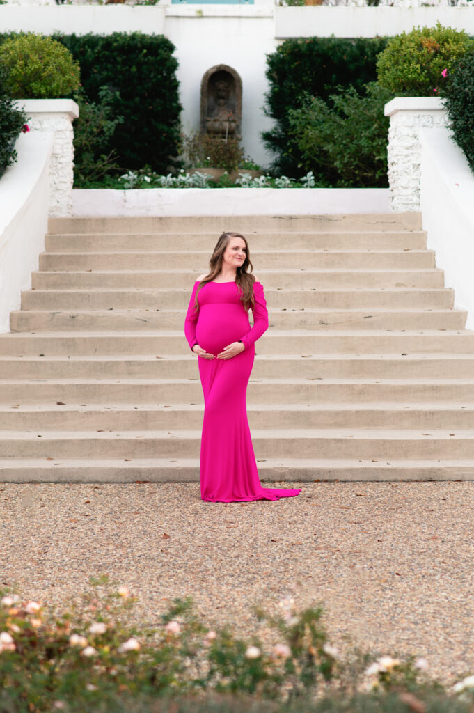 Pregnant woman in a form‑fitting bright pink gown stands barefoot on a gravel path in front of broad stone steps and manicured hedges, gently cradling her belly and looking off to the side.