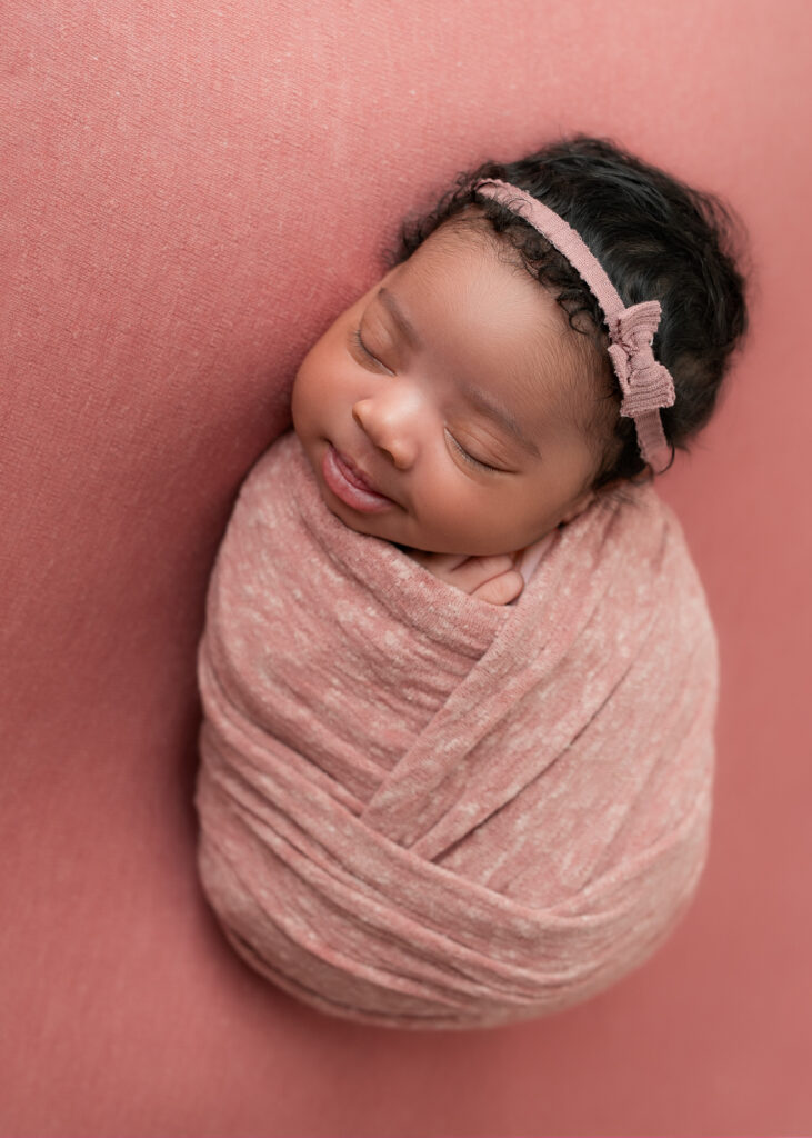 Newborn girl wrapped in blush knit, sleeping on soft pink backdrop
