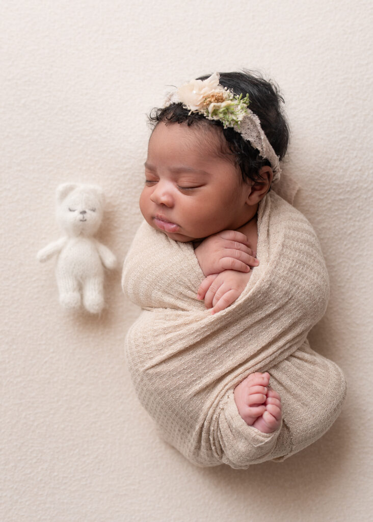 Baby swaddled in cream wrap, posed on white backdrop with teddy bear
