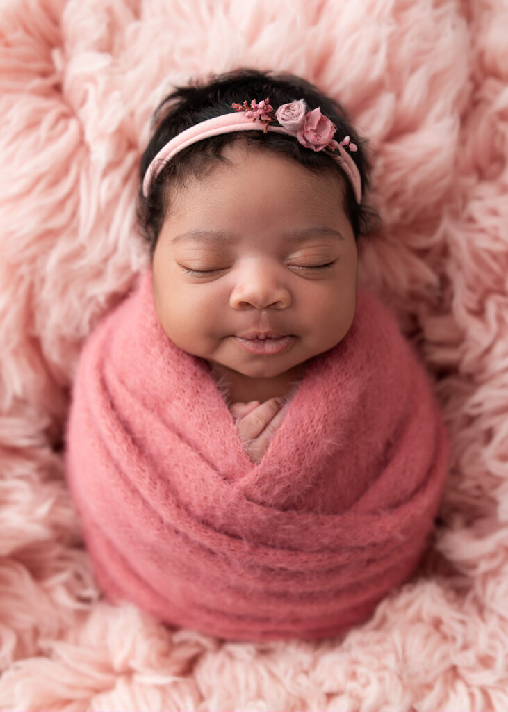 Girl baby swaddled in deep pink wrap with flower headband on peachy pink backdrop