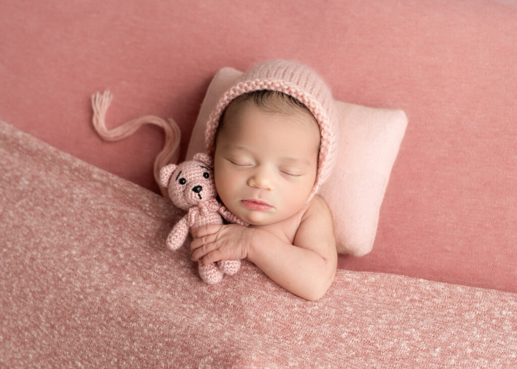 Girl baby in pale pink bonnet, holding tiny bear on warm blush backdrop
