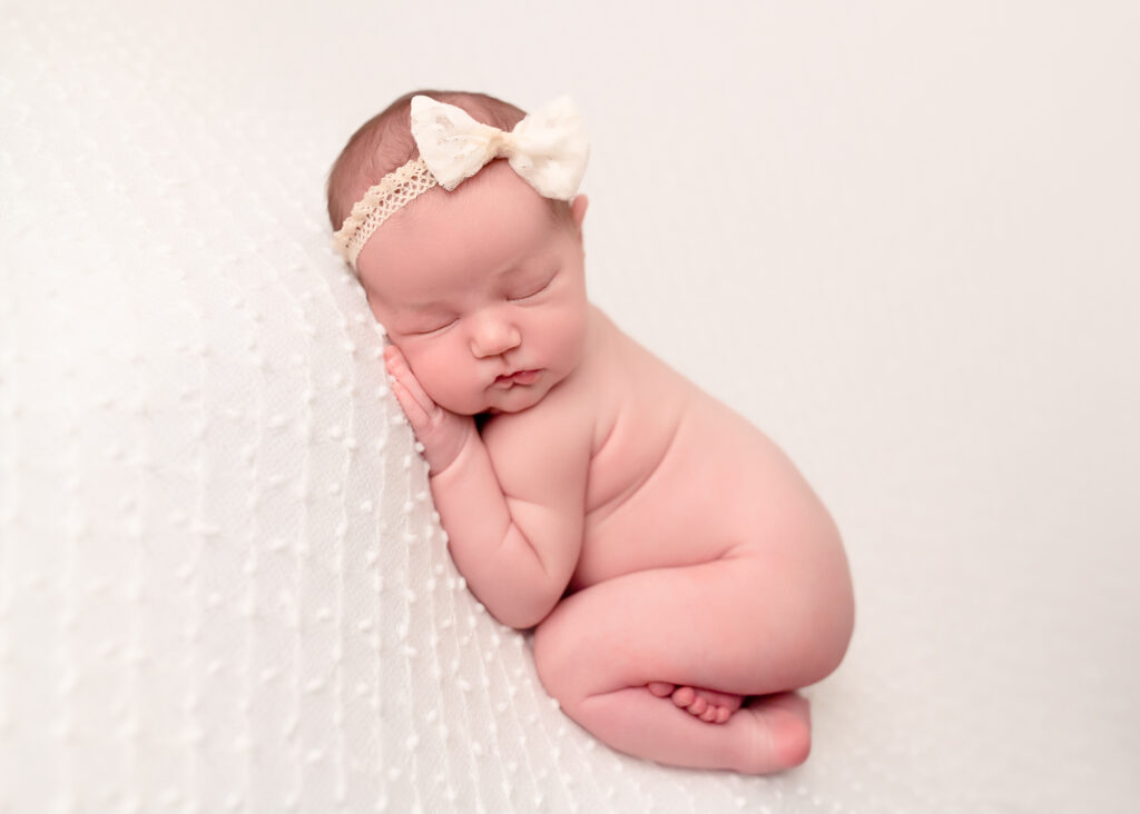 Nude-toned baby girl on white backdrop with ivory bow headband