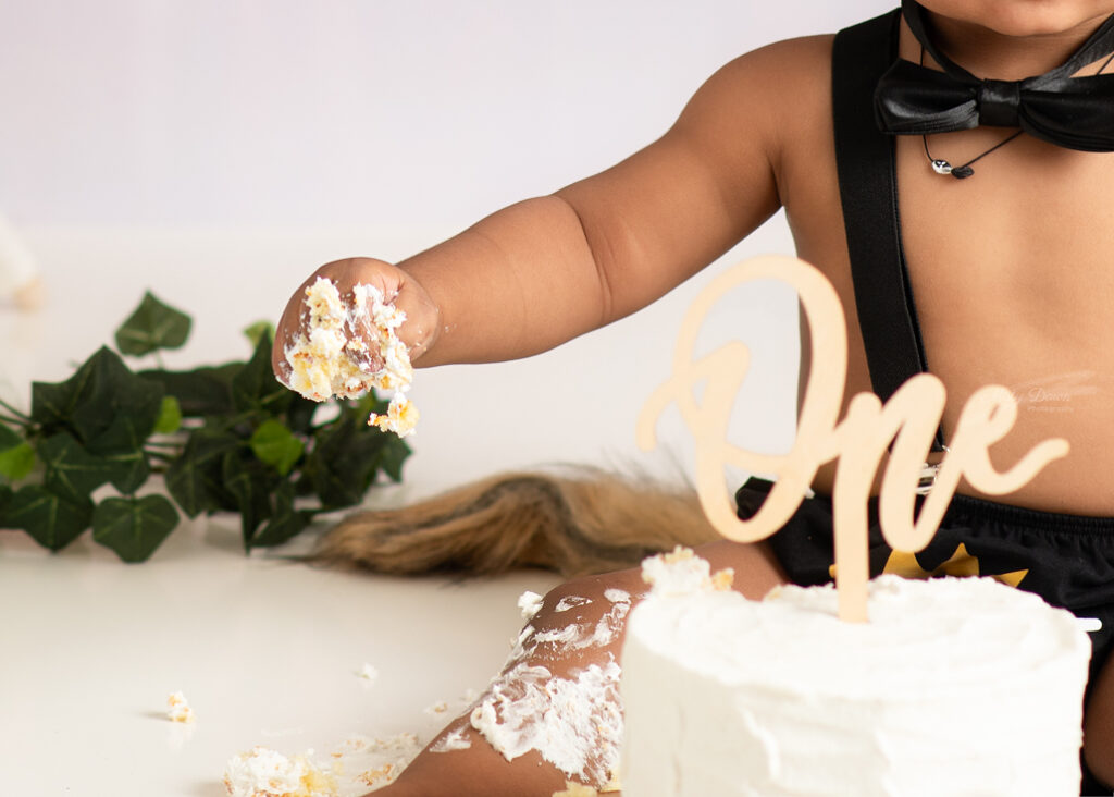Baby boy reaching for white cake with “One” topper during cake smash