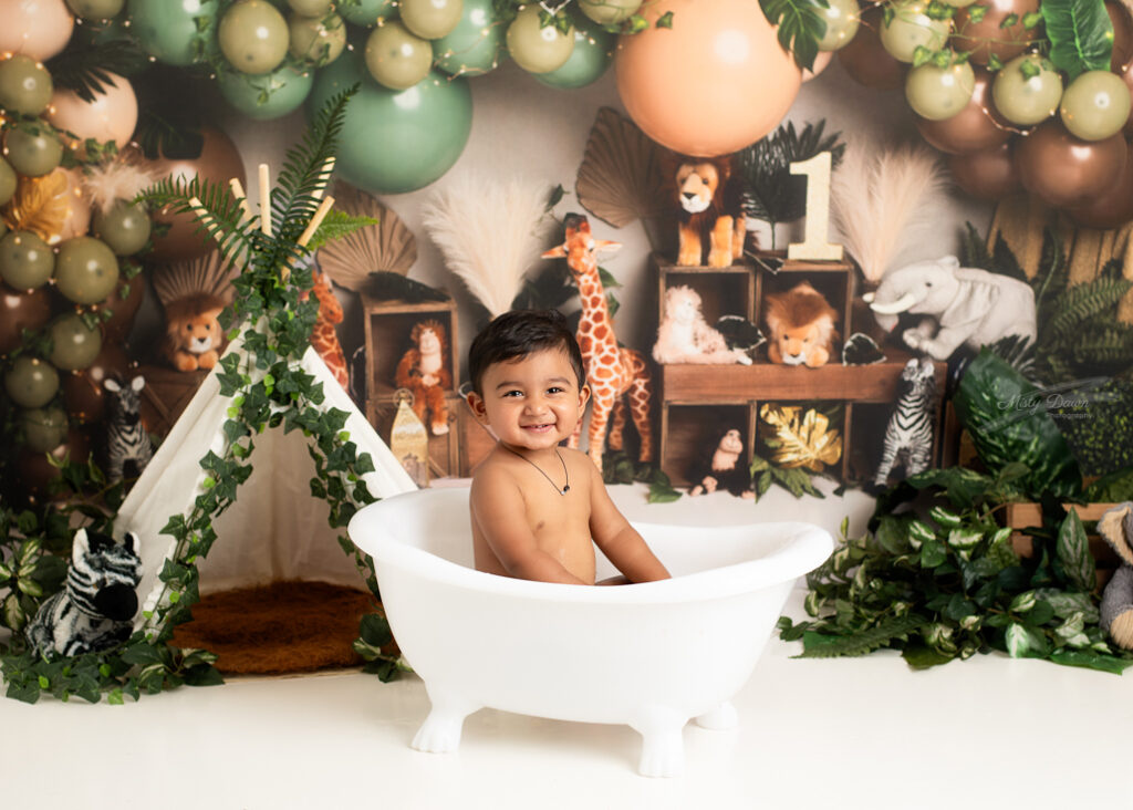 Smiling baby boy in white clawfoot tub during jungle-themed cake smash and splash session with animal backdrop and safari tent