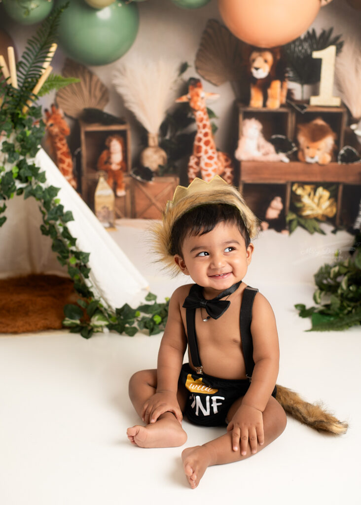 Smiling baby boy in black suspenders and crown during jungle-themed cake smash session