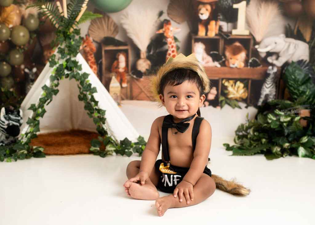 Baby boy sitting in front of tent and safari backdrop, smiling sweetly in first birthday outfit