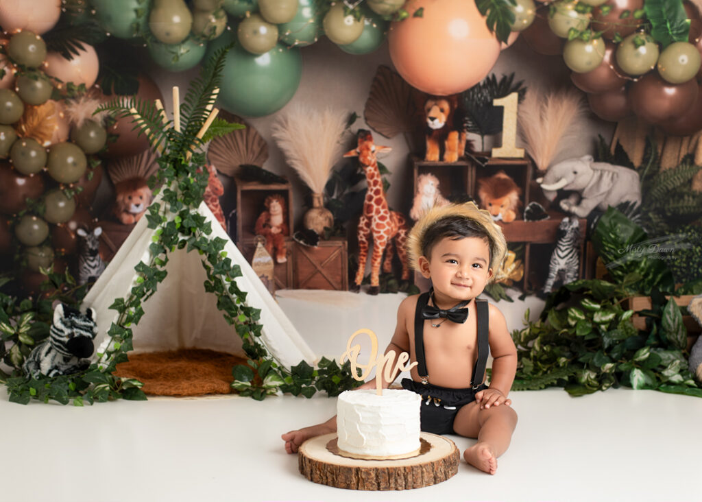 One-year-old boy in crown and bowtie sitting beside white cake during safari-themed first birthday photoshoot with plush animals and balloon garland