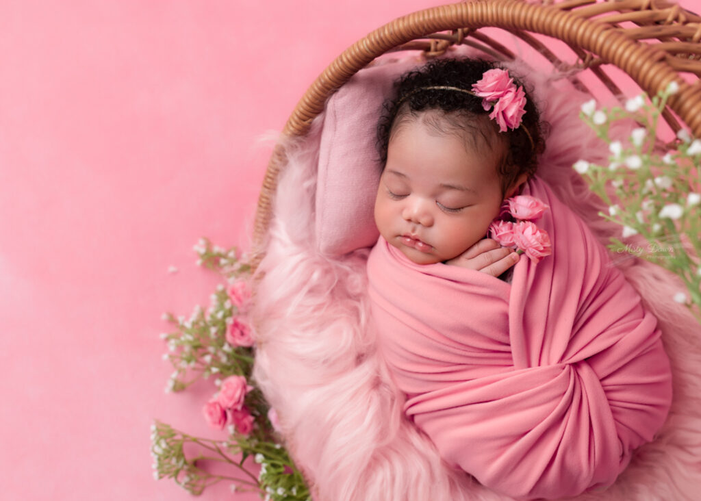 Newborn baby girl peacefully sleeping in a basket wrapped in pink swaddle with matching flowers, styled for a Birmingham newborn photography session by Misty Dawn Photography.