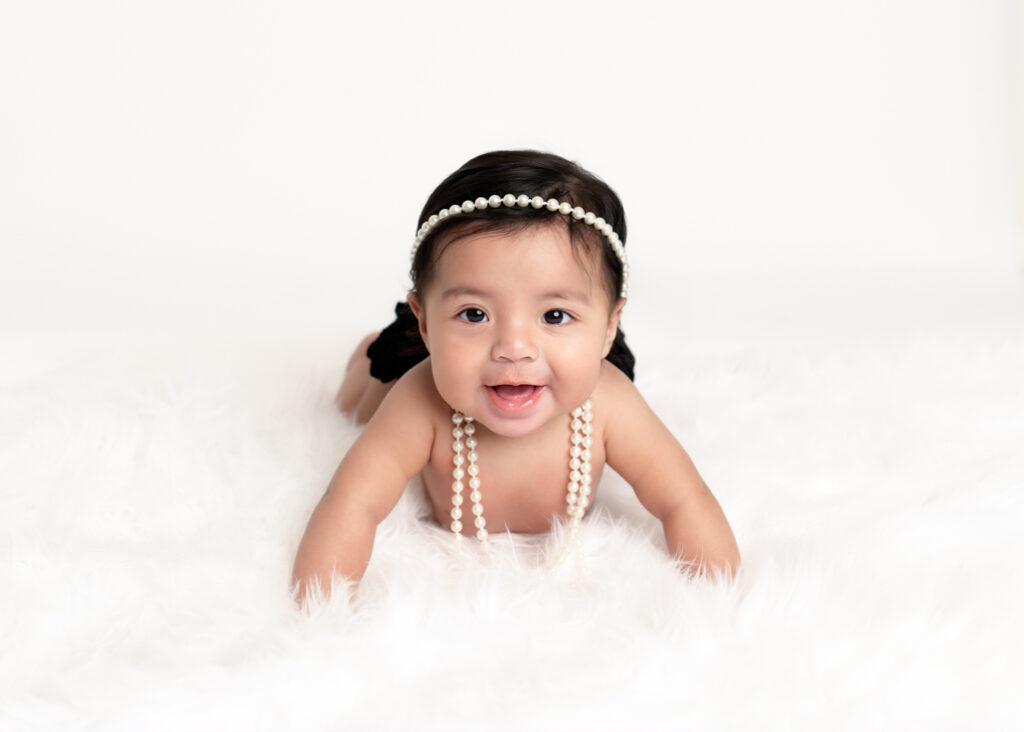 baby in crawling position on white fur, wearing black bloomers and white pearls during a Sitter session by Misty Dawn Photography in Birmingham.