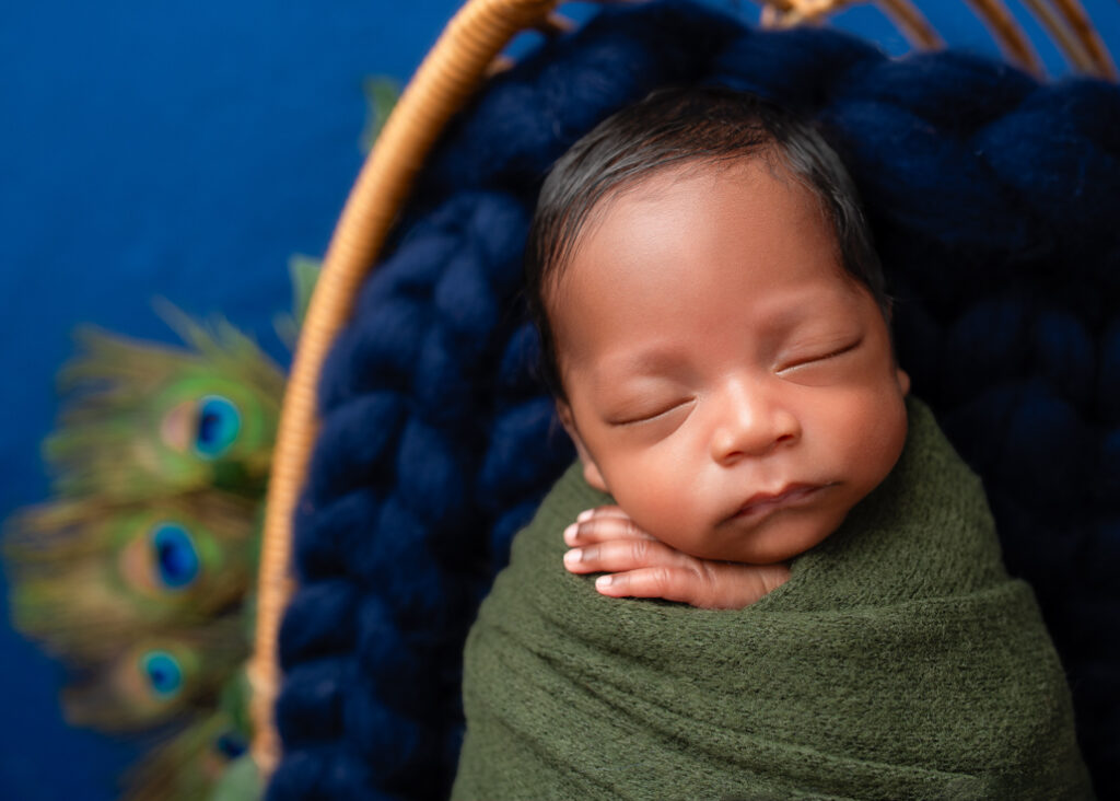 baby wrapped in green and sleeping in wicker basket and dark blue woven blanket with peacock feathers poking out from basket.