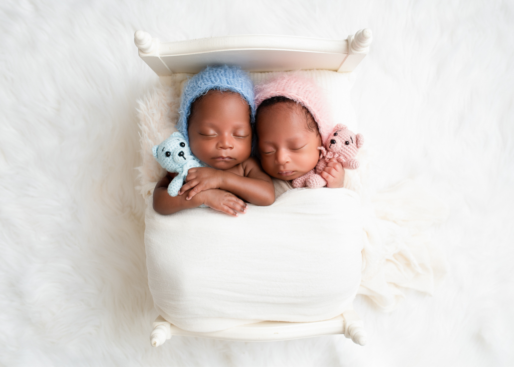 a boy and a girl twin newborns cuddled together wearing blue and pink on a white bed and white background