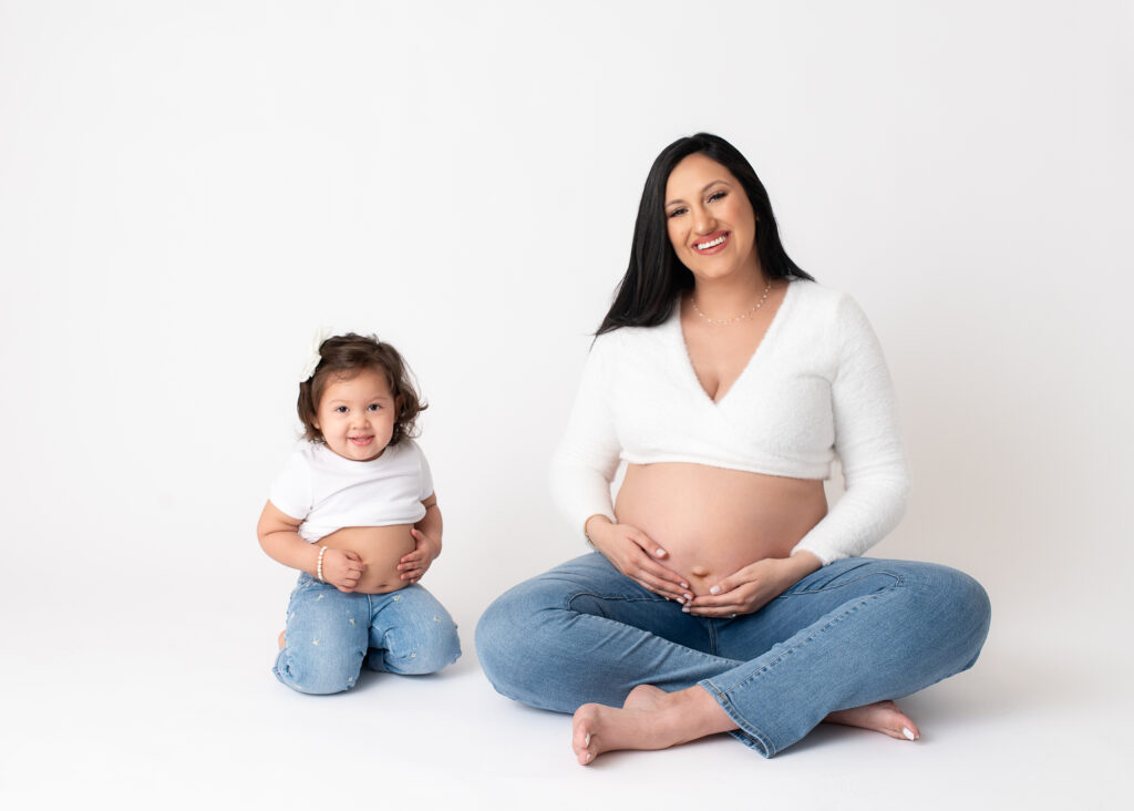 Pregnant mother and her 1 year old baby girl holding their bellies while wearing white shirt and blue jeans on a white background during a Maternity with family session at Misty Dawn Photography in Birmingham.