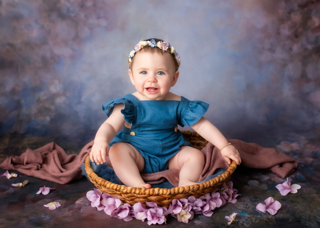 Smiling baby girl sitting in a wicker basket with a floral headband and blue romper, surrounded by soft pink flower petals during a sitter session by Misty Dawn Photography in Birmingham.