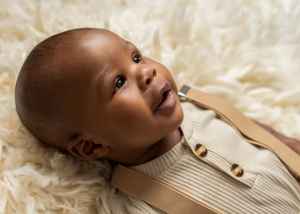 Adorable baby boy lying on a soft white fur blanket, wearing a beige outfit with suspenders, captured during a milestone photography session in Birmingham by Misty Dawn Photography