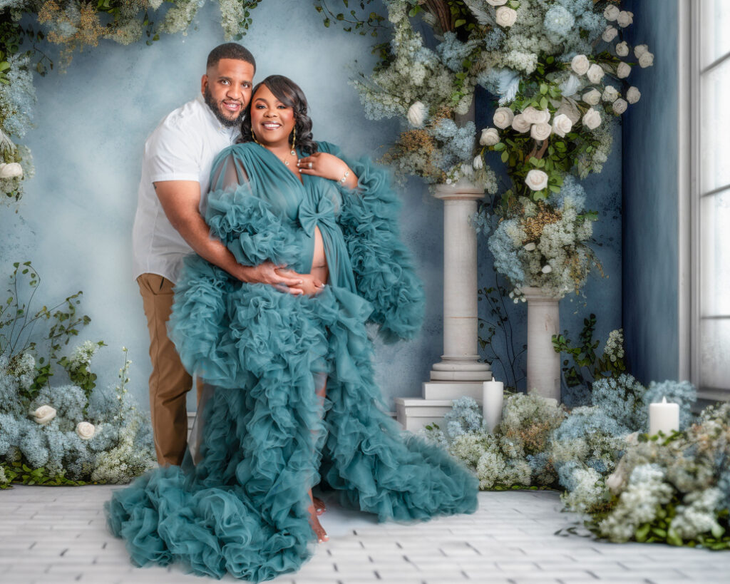 Expecting couple posing in an elegant floral studio with a dreamy blue and white theme, featuring the mother in a flowing teal maternity gown