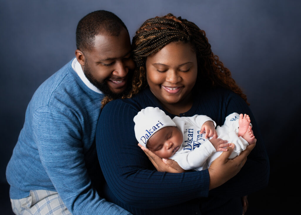 A new mother and father dressed in blue with a lighter blue background holding boy baby dressed in white with his name in blue