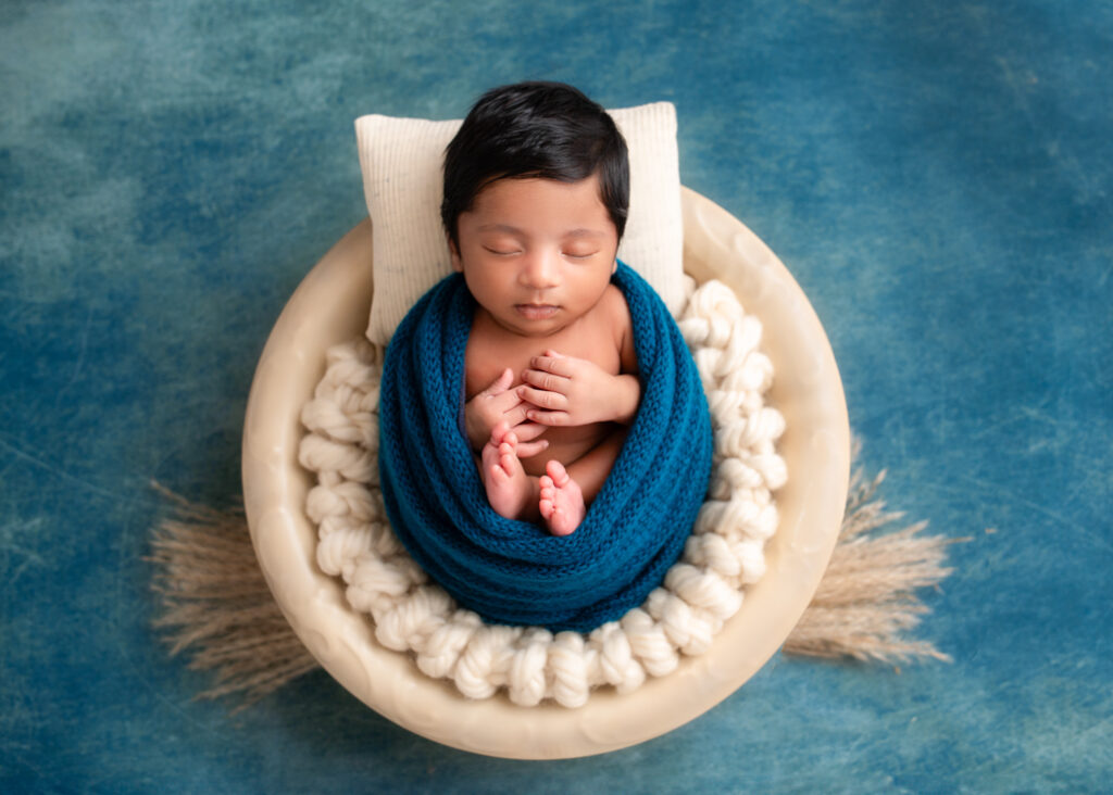 a newborn picture in Alabama that is wrapped in blue with only hands and feet exposed laying in a bowl with pillow and a blue background