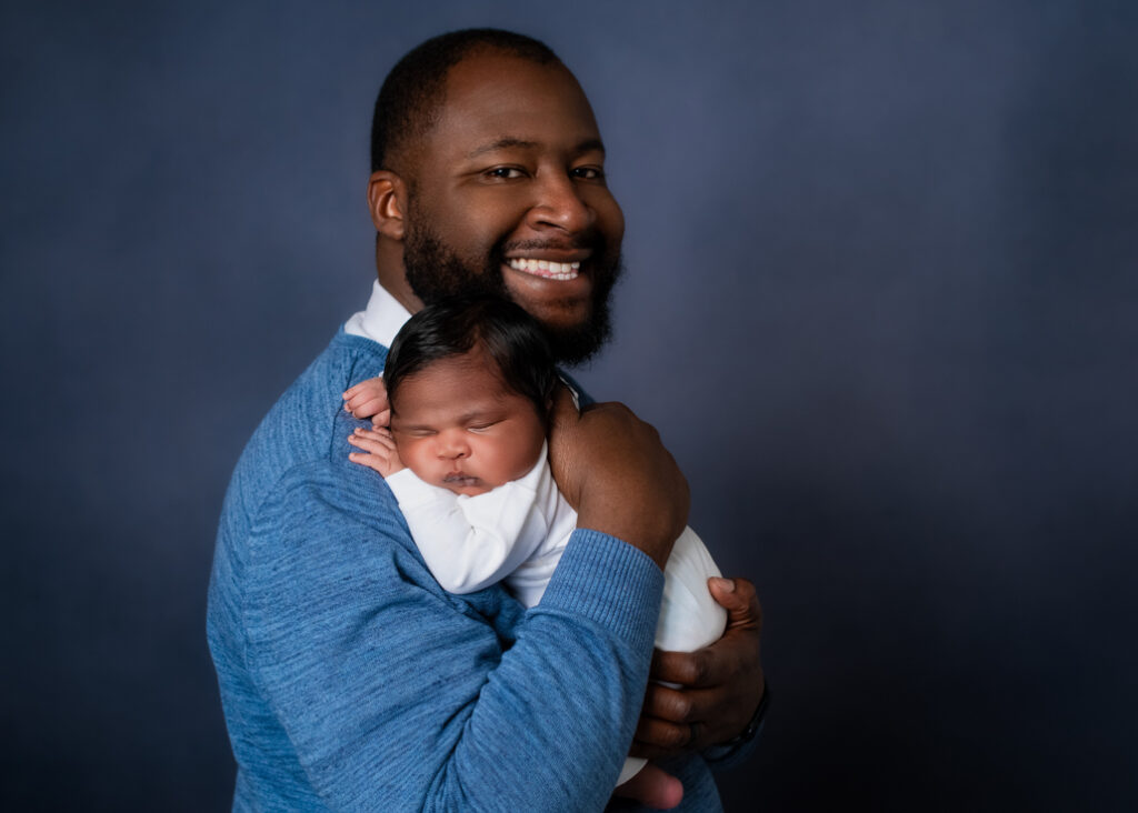 A new father dressed in blue with a lighter blue background holding her baby boy dressed in white with his name in blue