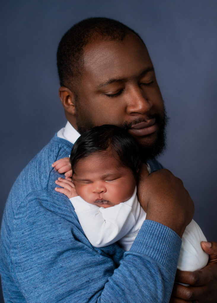 A new father dressed in blue with a lighter blue background holding his baby boy dressed in white with his name in blue