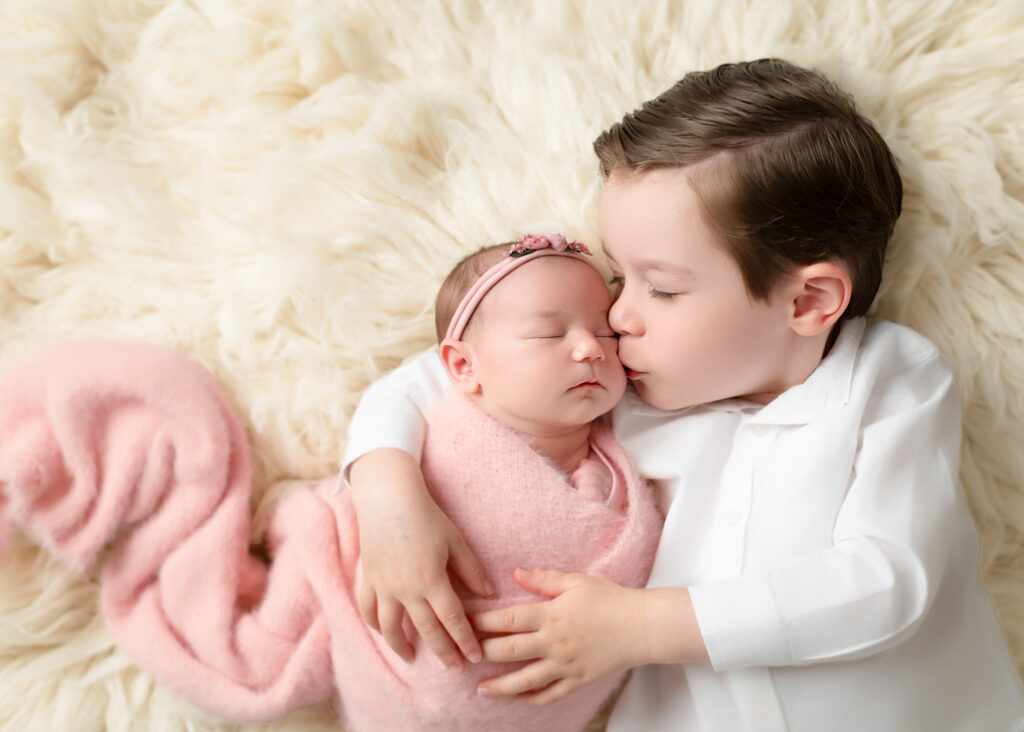 young boy dressed in a white shirt laying with his newborn sister while giving her a kiss. Newborn is wrapped in pink in this newborn portrait in Alabama