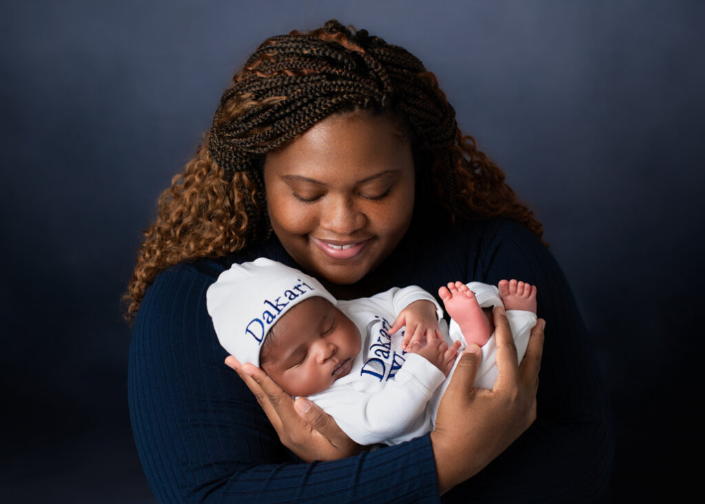 A new mother dressed in dark blue with a lighter blue background holding her baby boy dressed in white with his name in blue