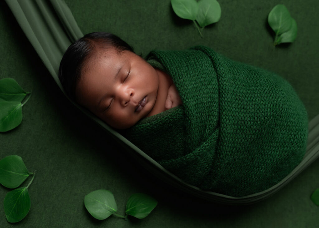 baby sleeping on green fabric and green hammock, wrapped in green with a leaves surrounding him