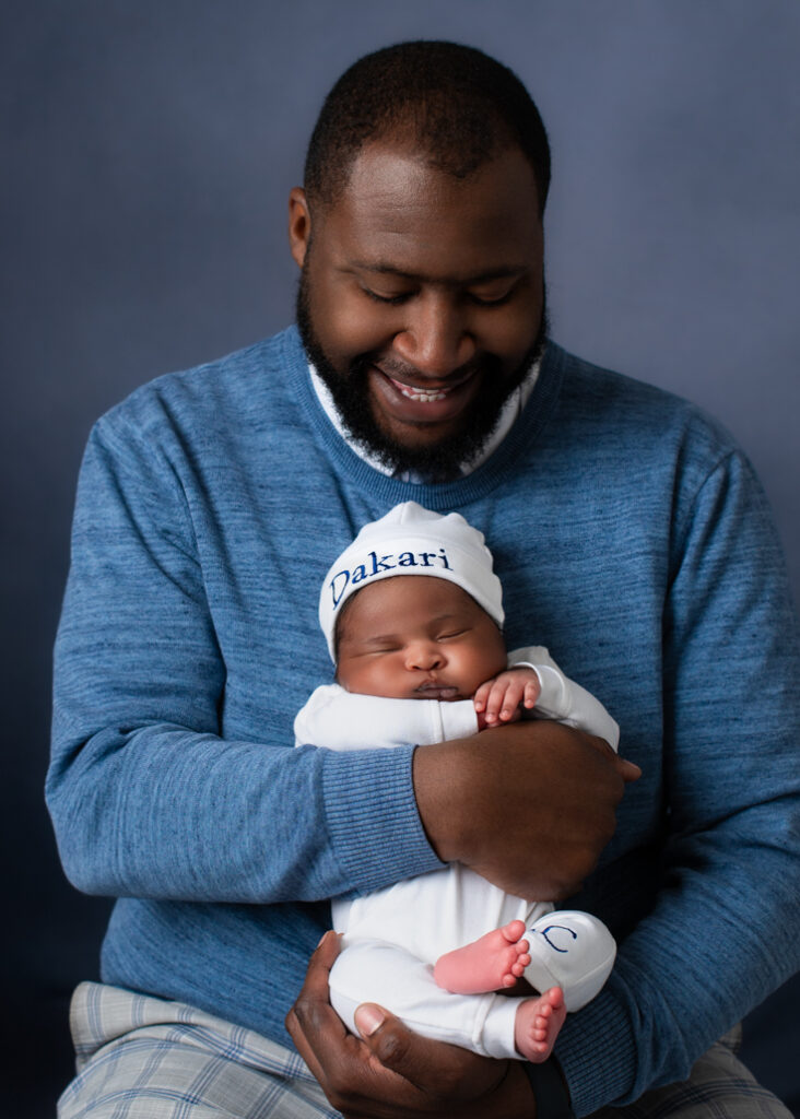 A new father dressed in blue with a lighter blue background holding his baby boy dressed in white with his name in blue