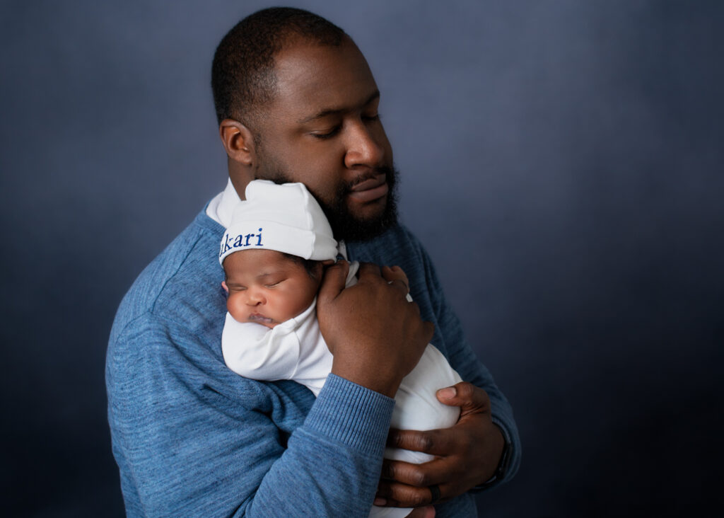 A new father dressed in blue with a lighter blue background holding his baby boy dressed in white with his name in blue