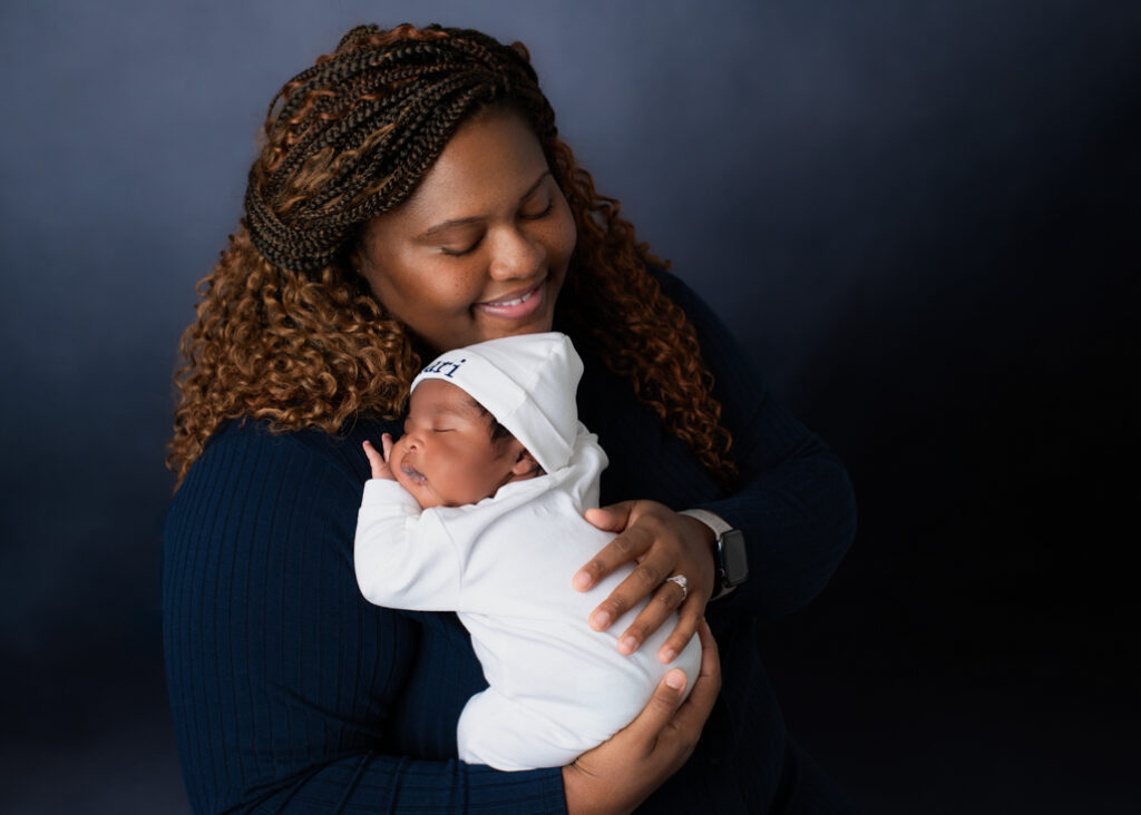 A new mother dressed in dark blue with a lighter blue background holding her baby boy dressed in white with his name in blue