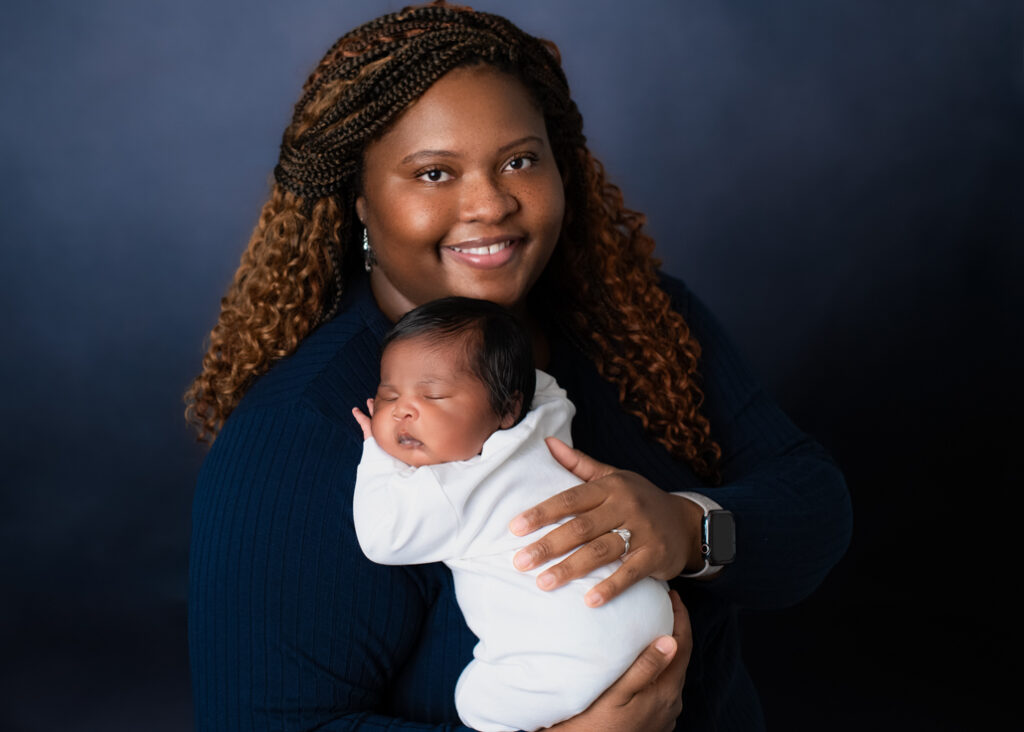 A new mother dressed in dark blue with a lighter blue background holding her baby boy dressed in white with his name in blue