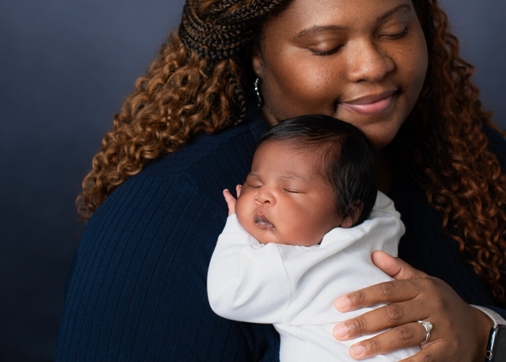 A new mother dressed in dark blue with a lighter blue background holding her baby boy dressed in white with his name in blue