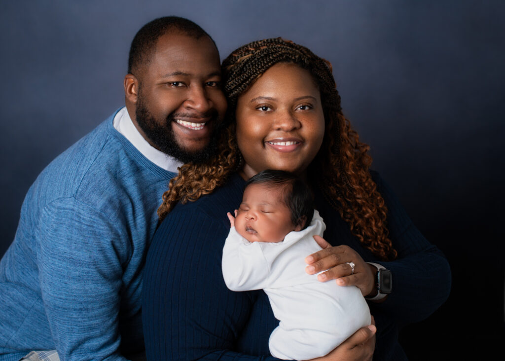 A new mother and father dressed in blue with a lighter blue background holding boy baby dressed in white with his name in blue