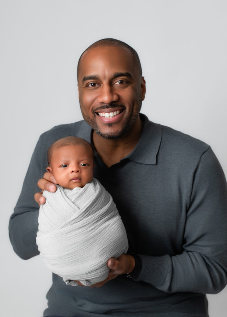 Twins' newborn photography featuring a proud father holding his swaddled newborn in a timeless and heartwarming portrait, celebrating the bond between father and child.