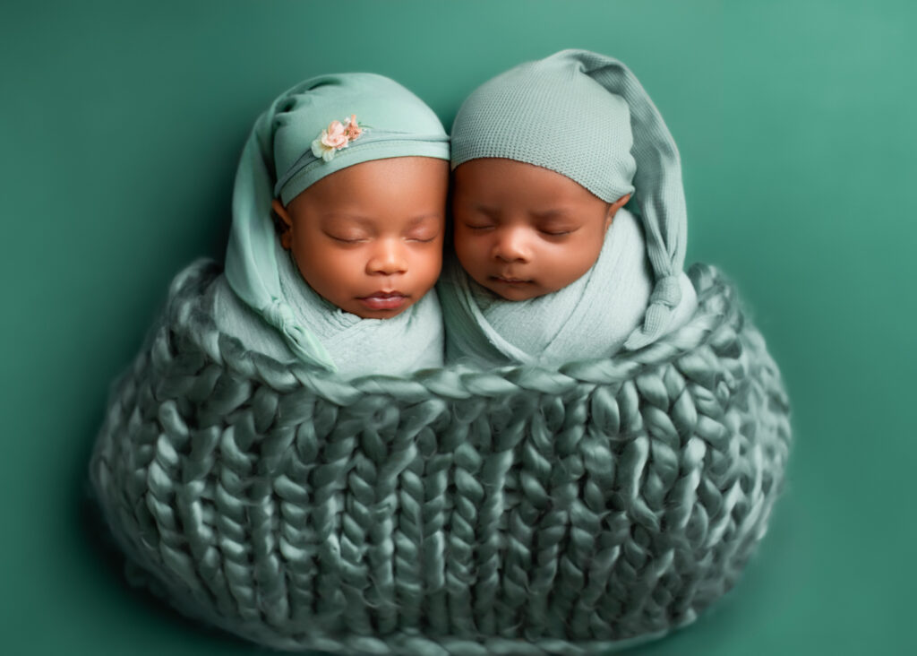 Twins' newborn photography featuring two peacefully sleeping newborn twins, swaddled in soft sage green wraps with matching hats. One twin wears a delicate floral headband, while the other dons a cozy knotted nightcap. They are nestled together in a chunky knit basket against a coordinating green backdrop, capturing the beauty of their serene expressions and perfect newborn features.