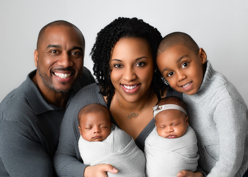 Twins' newborn photography featuring a joyful family portrait with parents and their older child embracing their newborn twins, both swaddled in soft gray wraps. A heartwarming moment celebrating the arrival of the twins