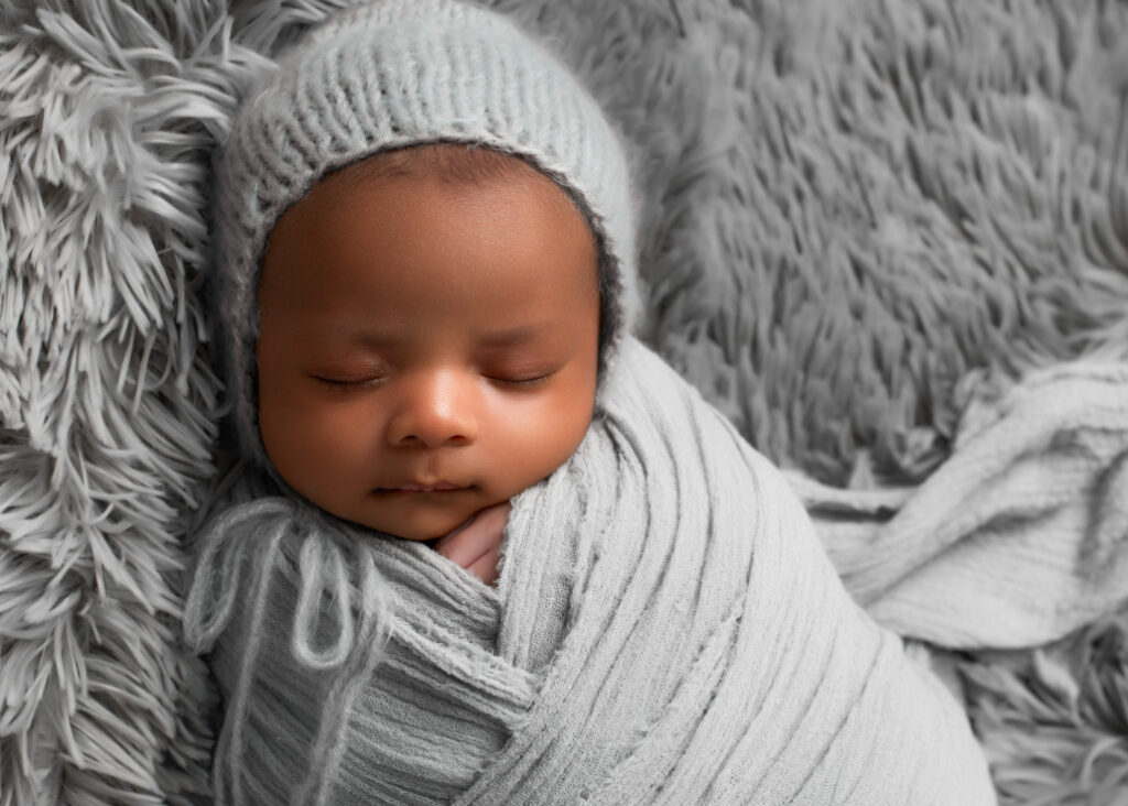 Twins' newborn photography highlighting a peaceful newborn baby boy, wrapped snugly in a soft gray swaddle and wearing a cozy knitted hat, resting on a textured gray blanket.