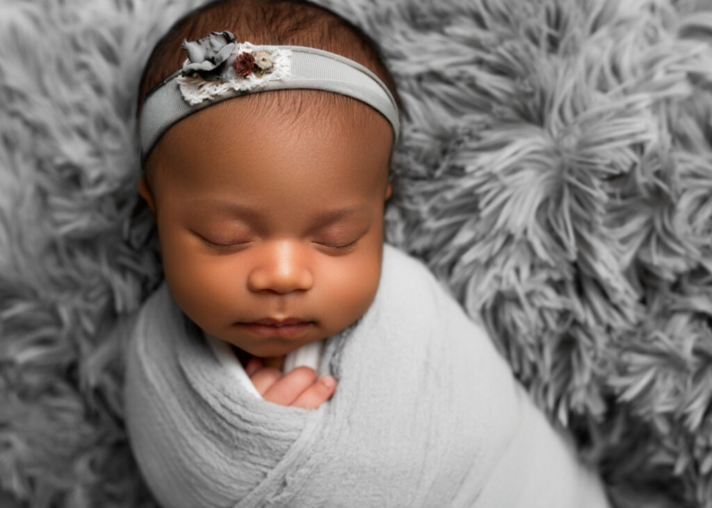 Twins' newborn photography capturing a serene close-up of a sleeping baby girl, swaddled in a soft gray wrap and adorned with a delicate floral headband, resting peacefully on a plush gray blanket