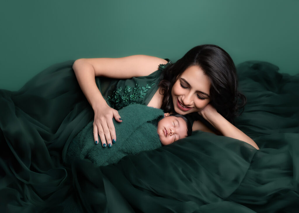 Mother in an emerald green gown lying next to her newborn wrapped in a matching green swaddle against a seamless green backdrop, showcasing fine-art styling, sculpted lighting, and intimate connection