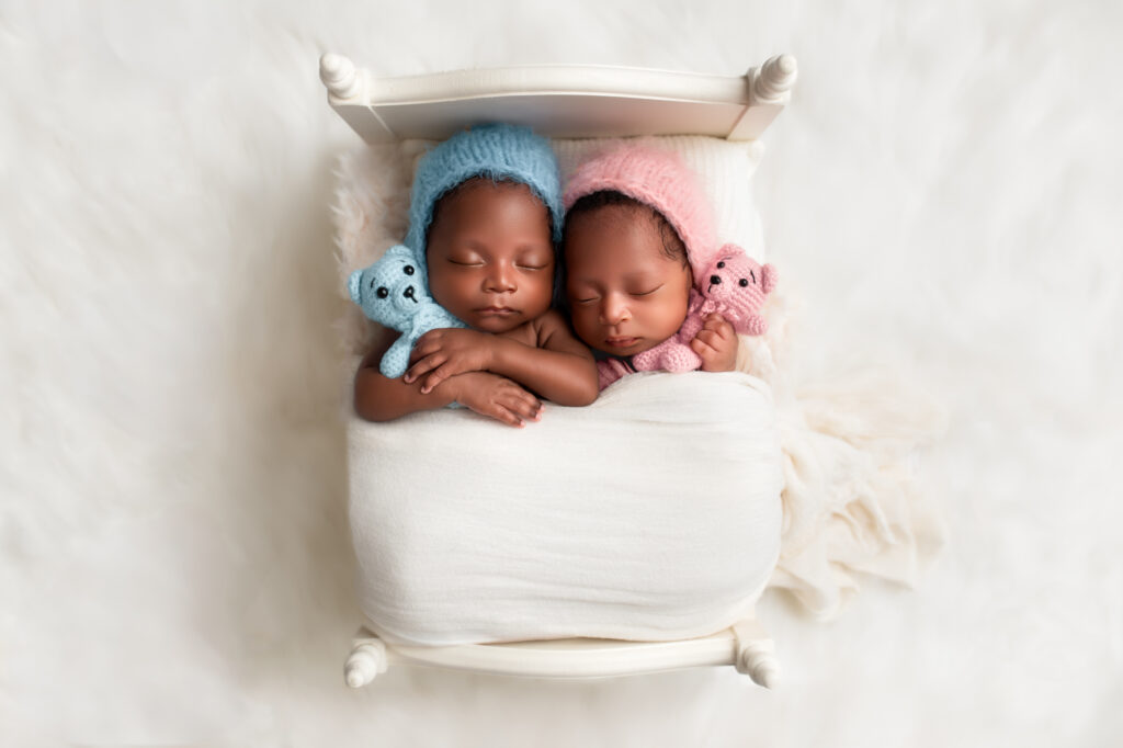 Twin newborns nestled side by side in a white miniature bed, each holding a pastel-colored teddy bear.