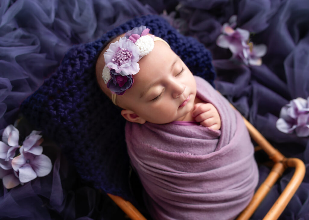 Newborn baby girl in lavender wrap with floral headband, softly lit in a rich purple-toned studio setup.